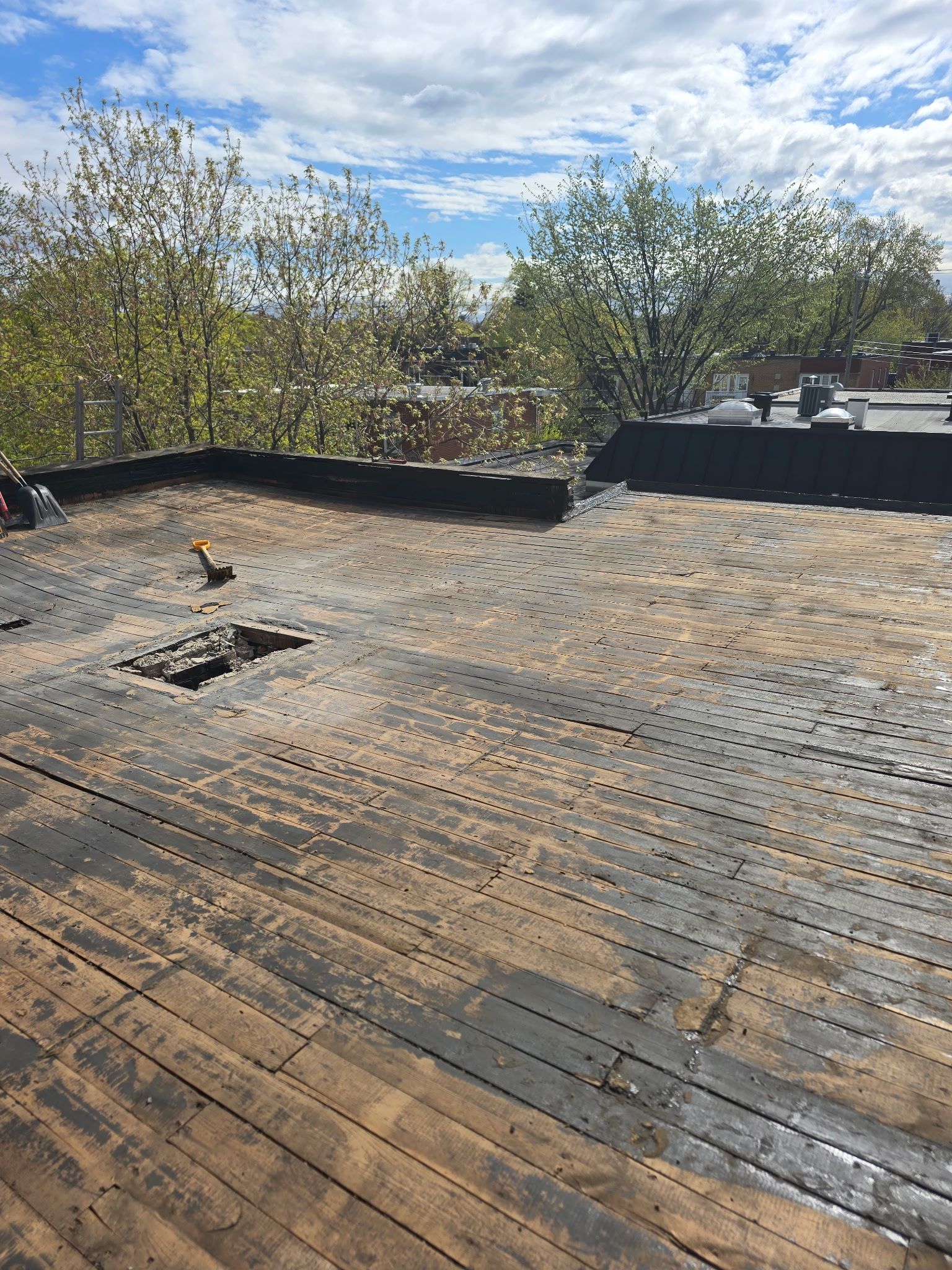 Toiture en construction avec platelage en bois apparent, bordure de toit noire et arbres sous un ciel partiellement nuageux