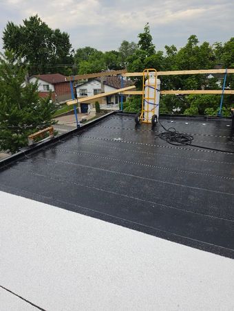 Toiture avec membrane d'étanchéité noire, rambardes de sécurité jaunes, arbres et maisons en arrière-plan