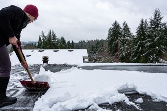 Une personne déneige un toit avec une pelle rouge, des arbres enneigés et une maison en arrière-plan.