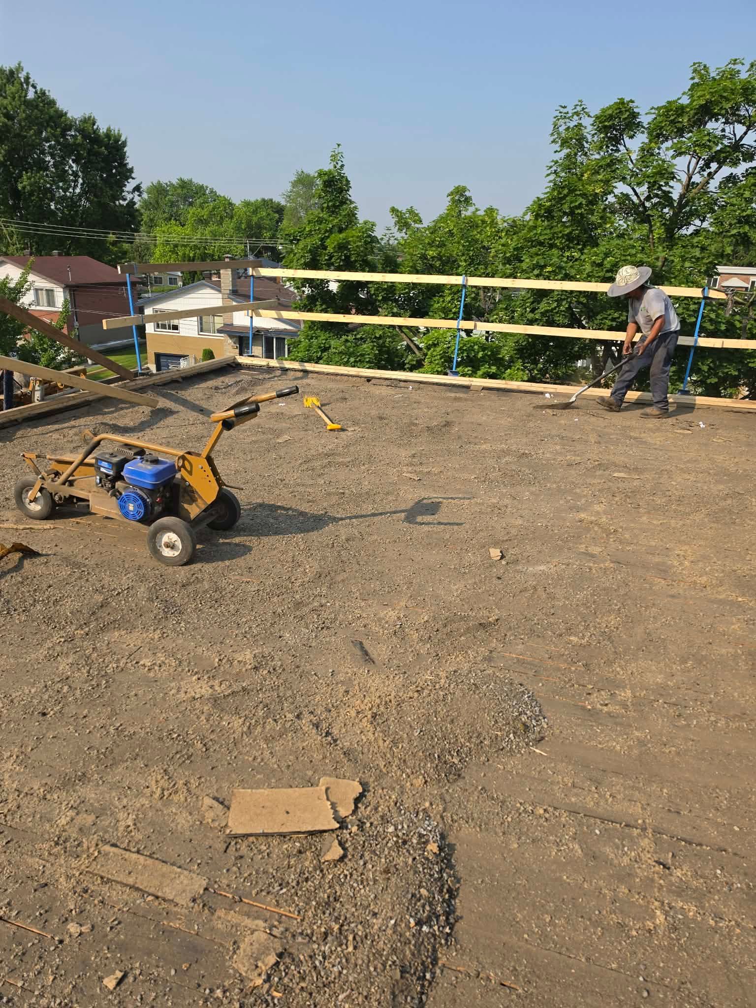Chantier de construction avec du gravier, des outils et un ouvrier installant des garde-corps jaunes sur un toit.