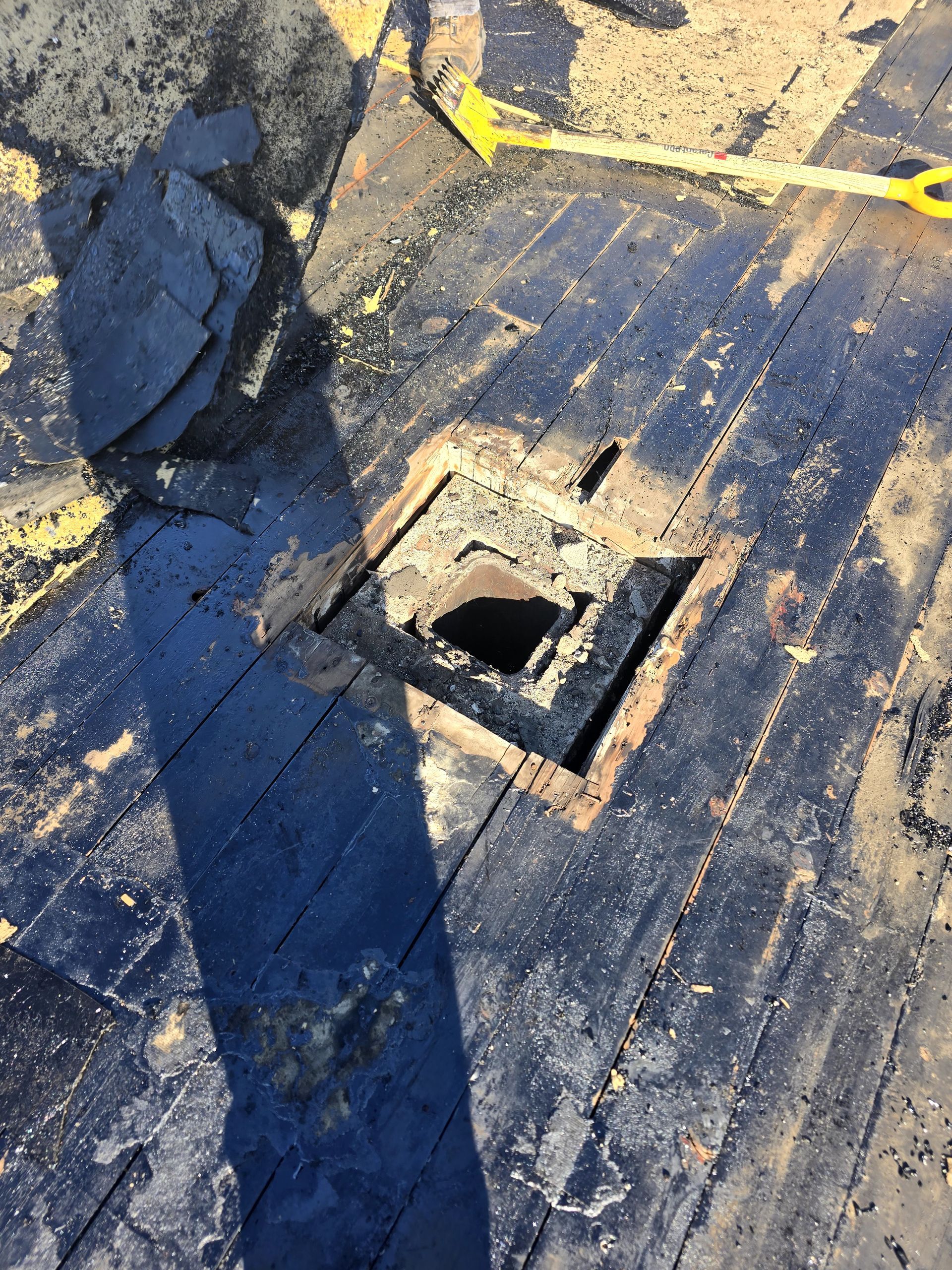 Ouverture carrée de ventilation de toit sur un toit noir vieilli par les intempéries, parsemé de débris et d'ombres dues au soleil.