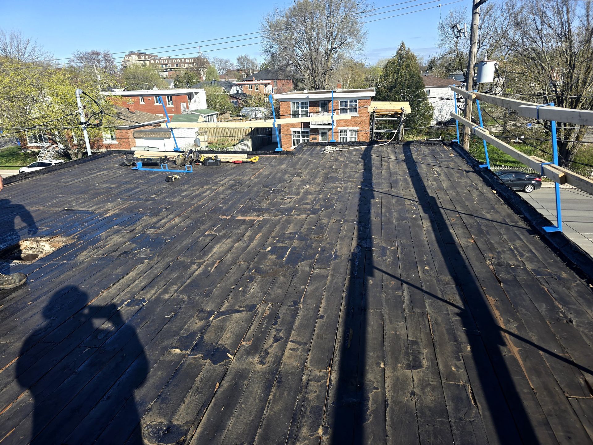 Toiture recouverte de feutre bitumé foncé, rambarde à droite et quartier résidentiel lumineux en arrière-plan.
