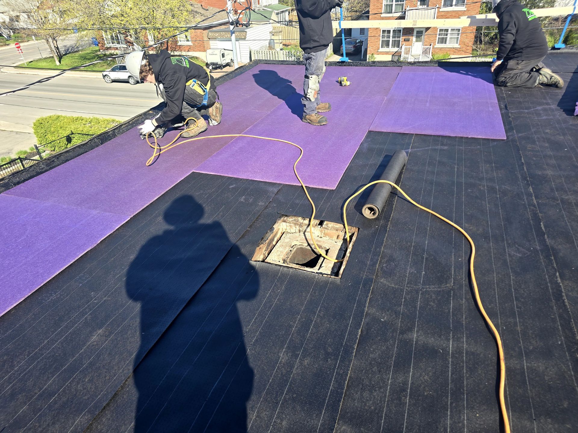 Des ouvriers installent une toiture violette sur un toit plat, laissant apparaître une ouverture de puits de lumière et une sous-couche noire.
