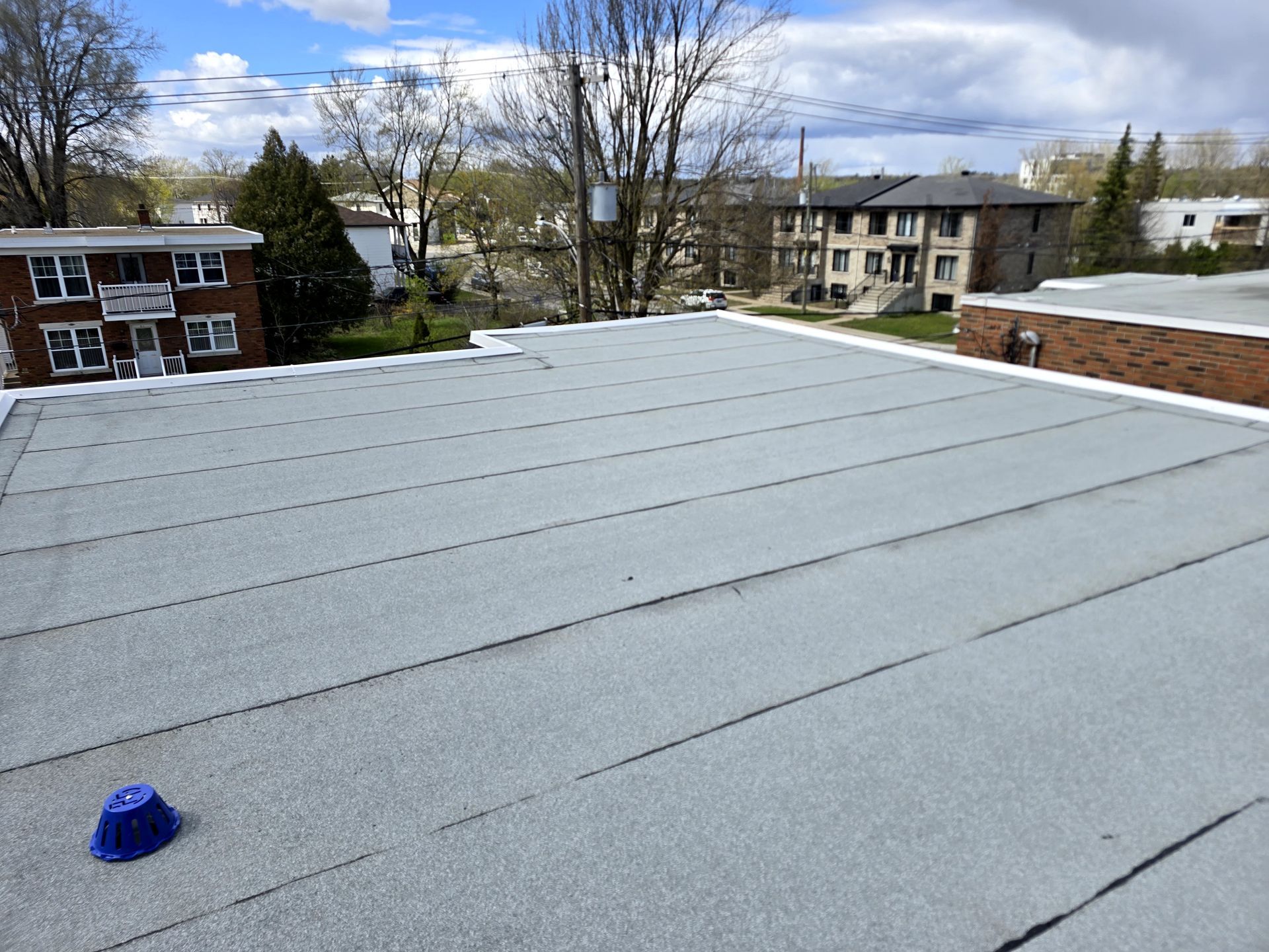 Toiture recouverte d'une membrane grise, avec un petit objet bleu au premier plan, et des bâtiments et des arbres à l'arrière-plan.