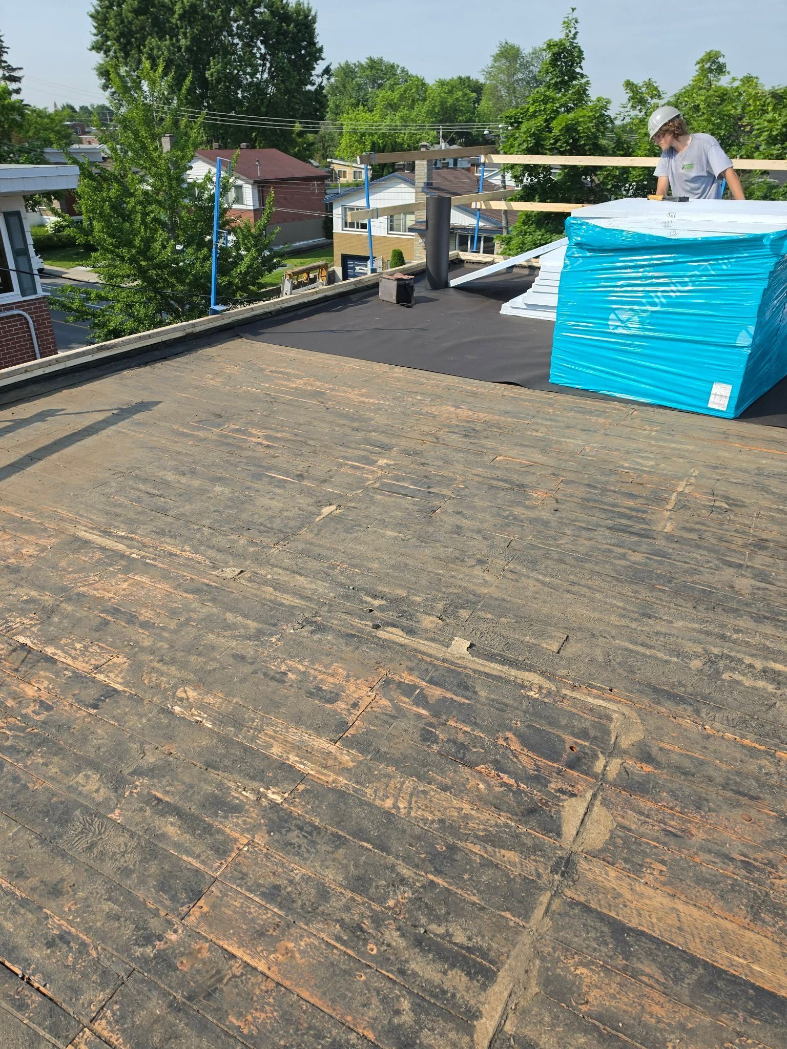 Chantier sur un toit avec une grande surface noire, des matériaux emballés en bleu et des ouvriers près du bord sous un ciel dégagé