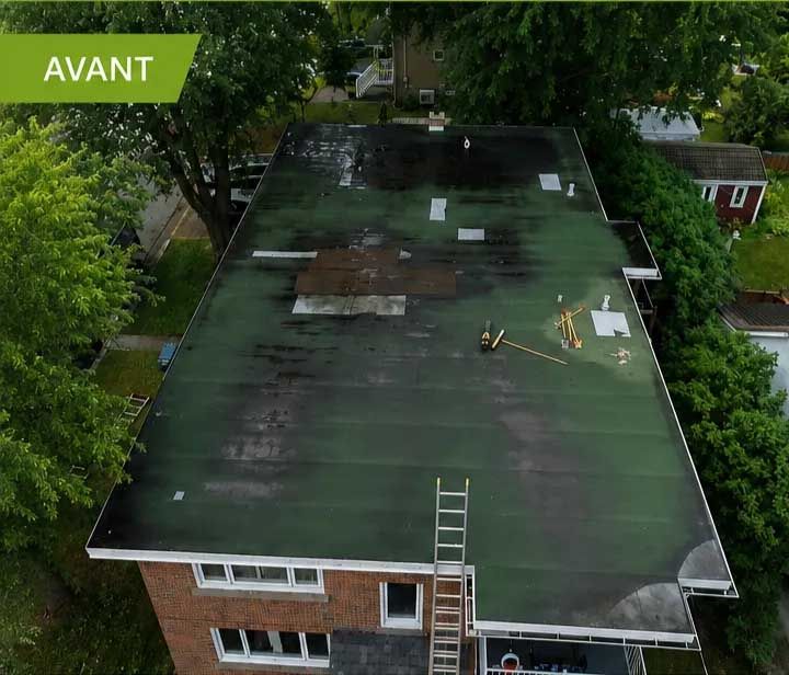 Vue aérienne d'un toit plat recouvert d'un revêtement vert, présentant des zones sombres, et d'un ouvrier près d'une échelle ; des arbres et des maisons se trouvent à proximité.