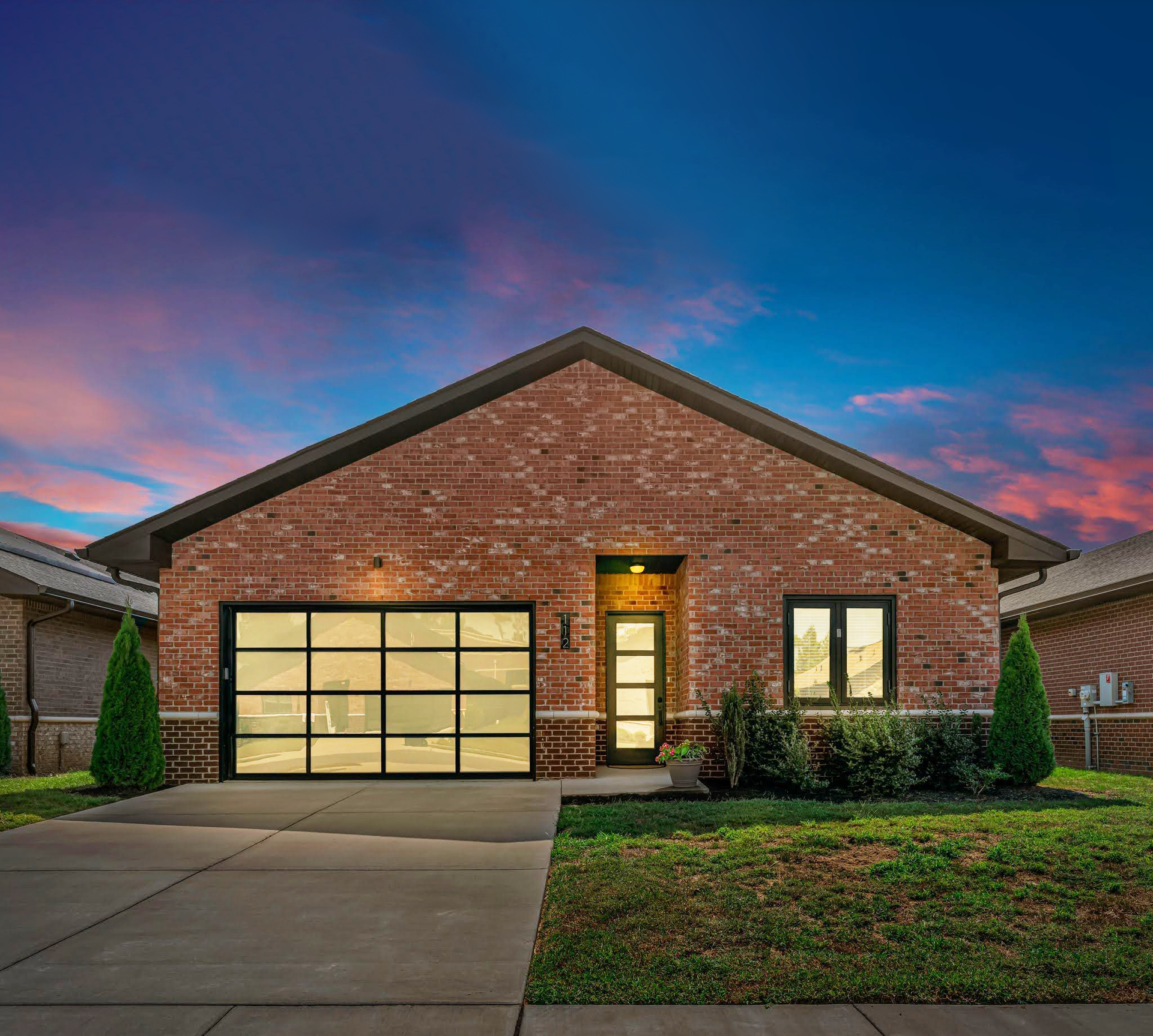Brick single-story house with two-car garage and lit front door at dusk under a pink-blue sky