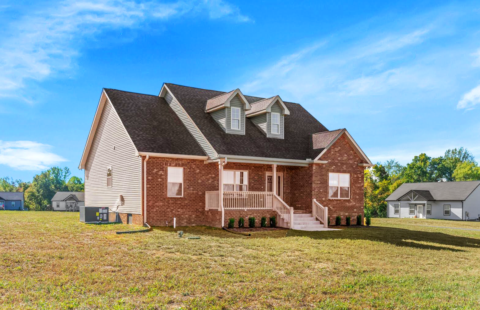 Large brick house with a front porch and dormer windows on a sunny grassy lot under a blue sky