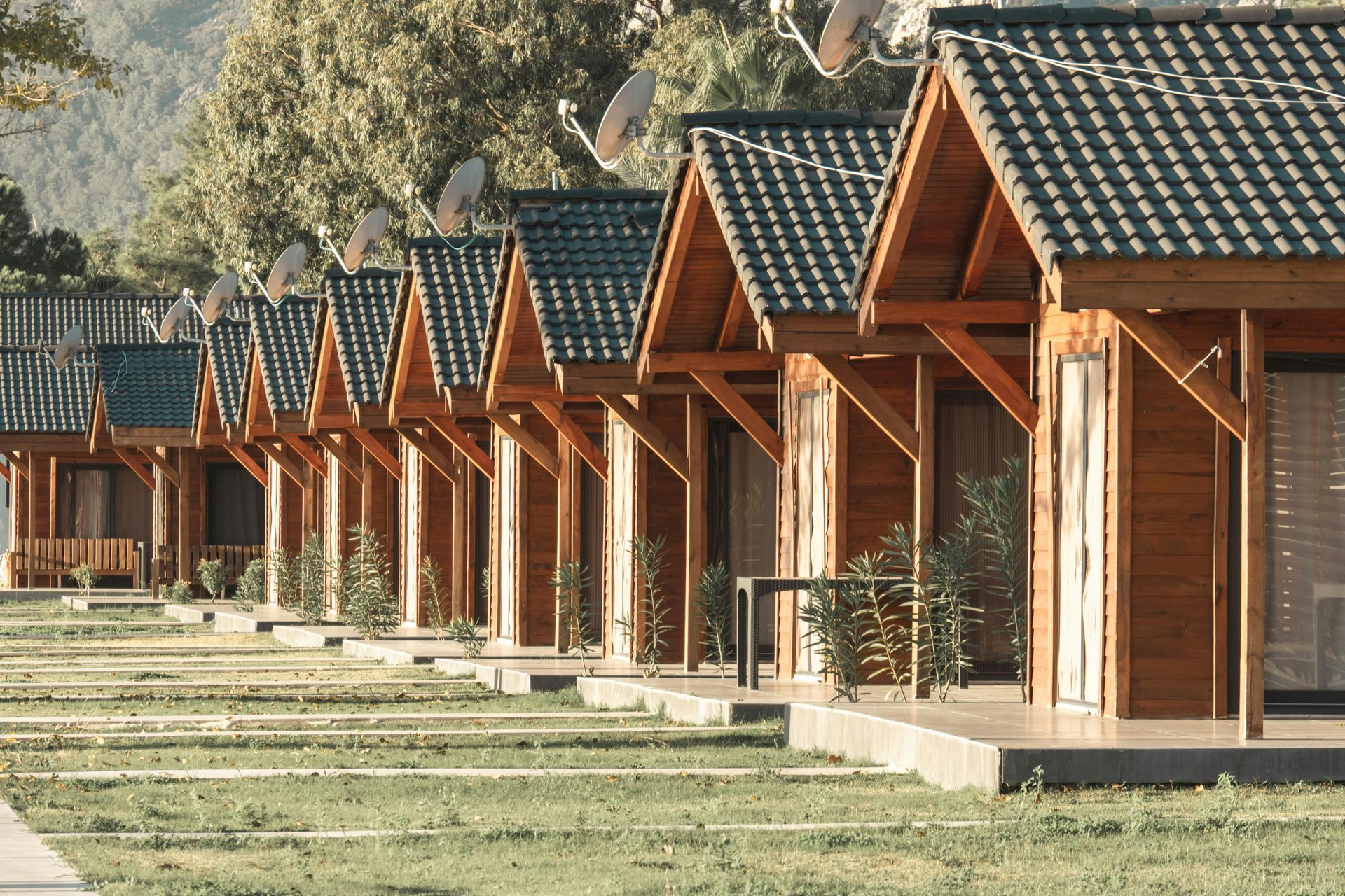 Row of small wooden cabins with pitched roofs beside a grassy path in a sunny park-like setting