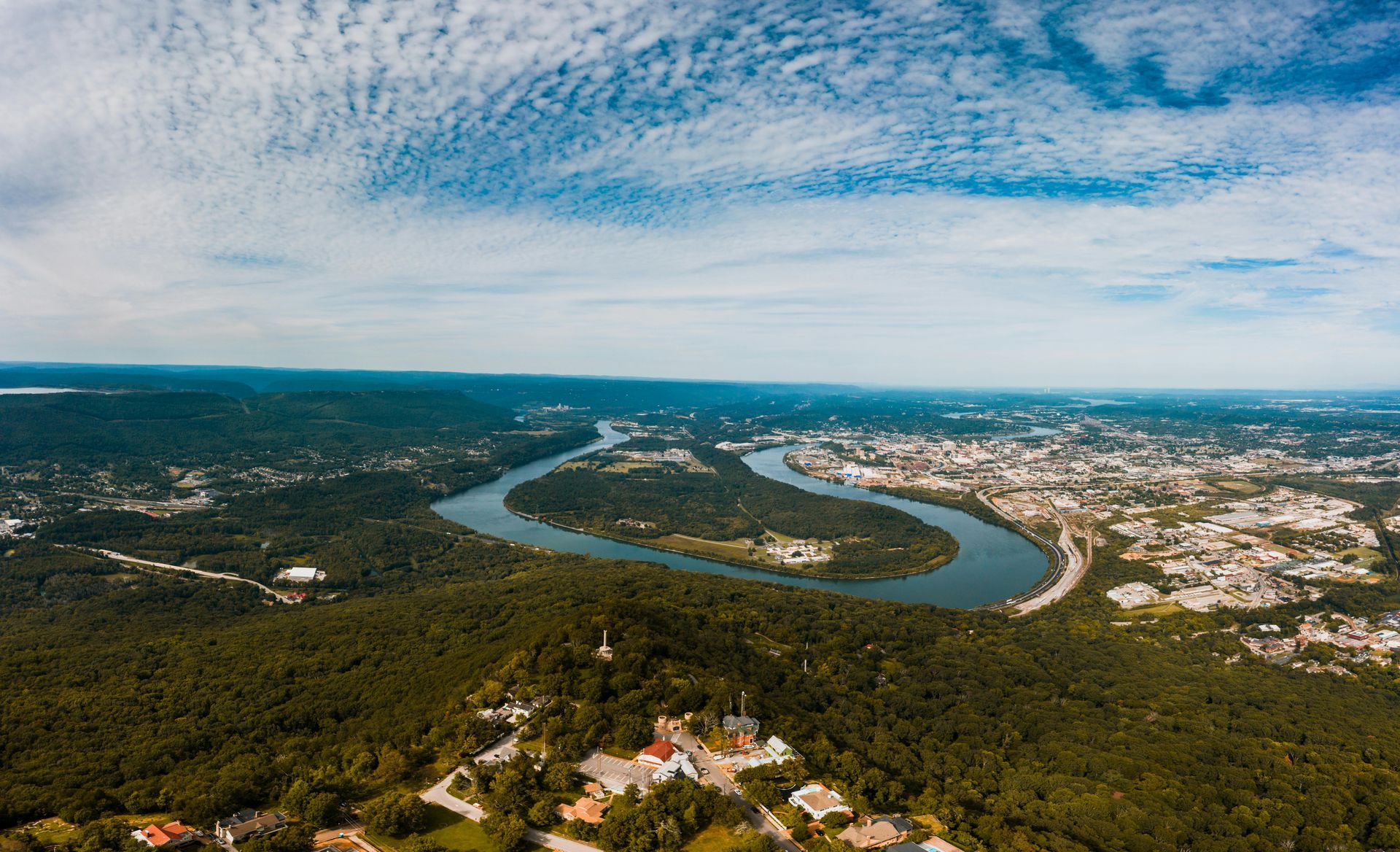 Aerial view of a winding river through green forest beside a sprawling town under a cloudy blue sky