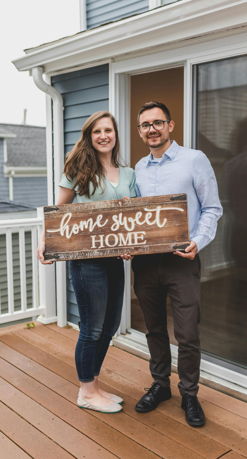 Couple standing on a porch holding a wooden sign that says “Home Sweet Home”