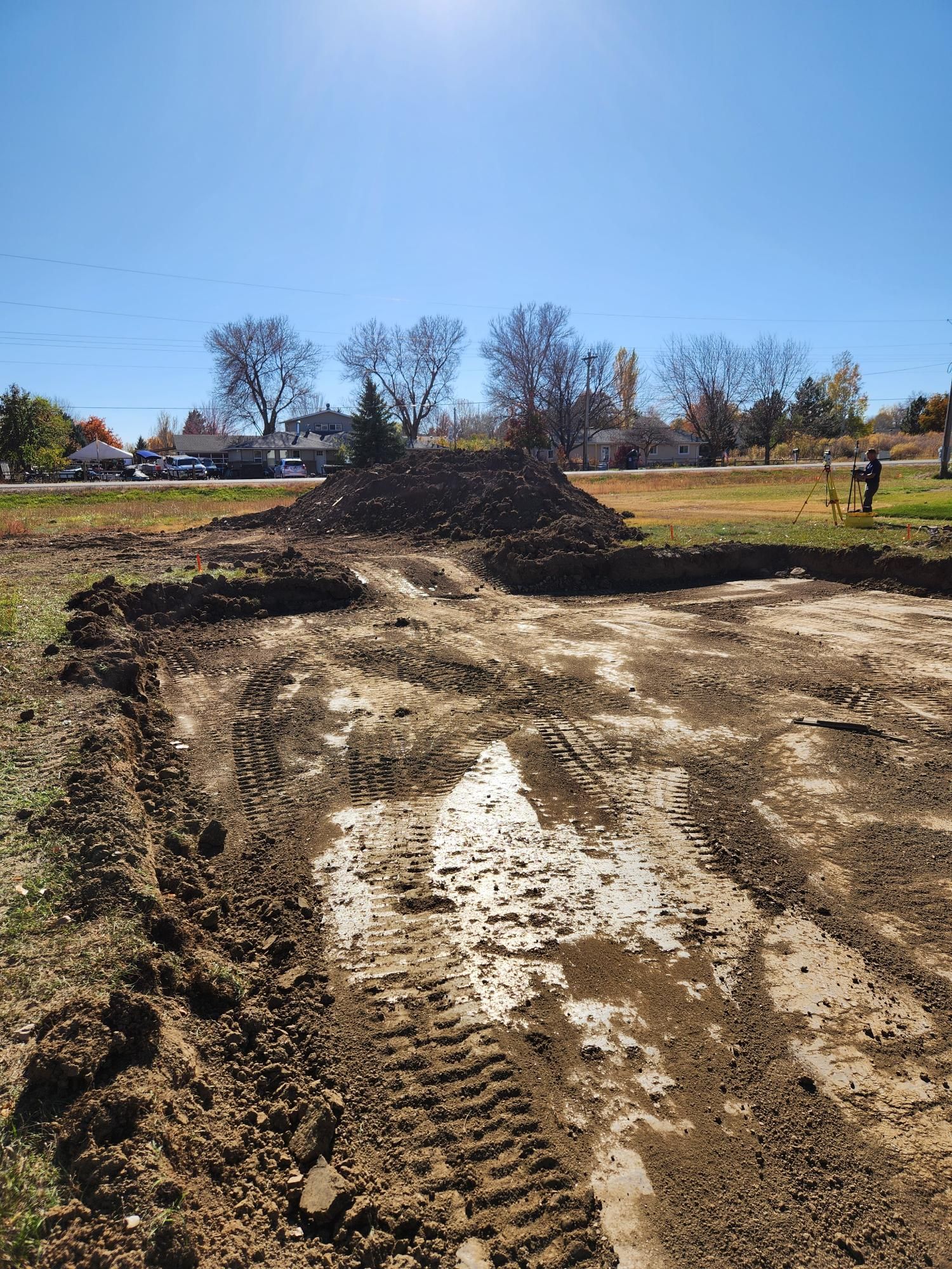 Muddy dirt road with tire tracks and puddles beside a field under a clear blue sky, with a person in the distance