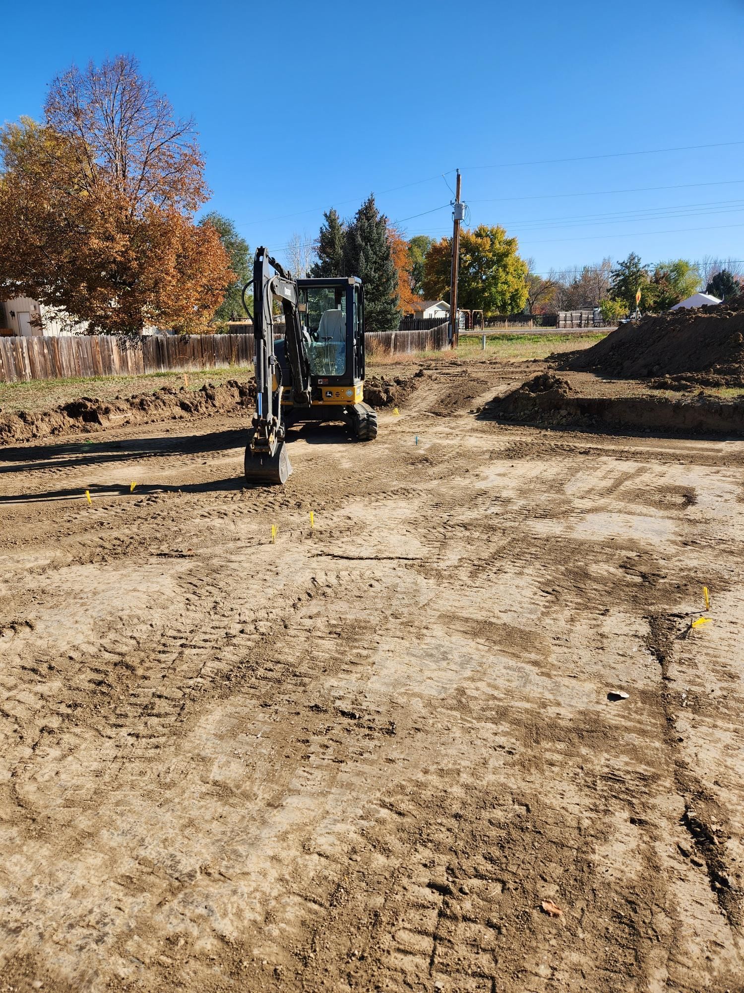 Excavator at a dirt construction site under a clear blue sky, with trees and piles of earth nearby