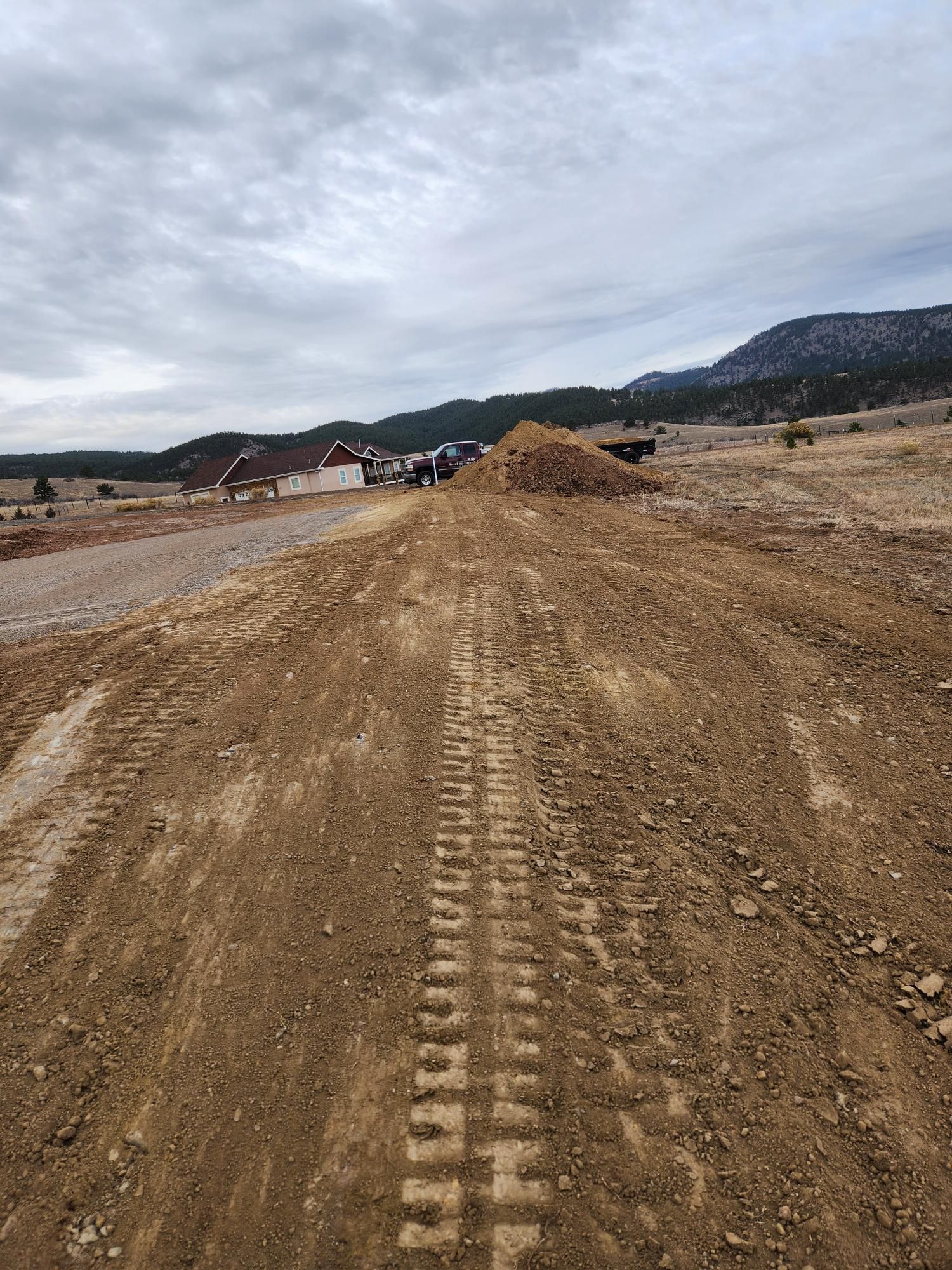 Dirt road under construction leading toward houses and hills beneath a cloudy sky
