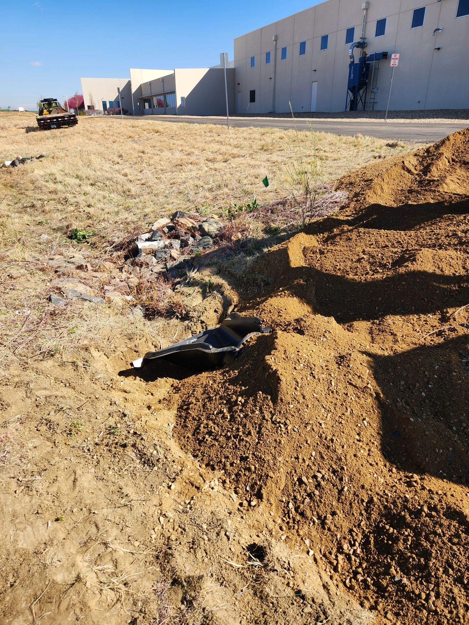 Dirt lot beside a white industrial building, with a small excavator and piles of soil in the foreground.
