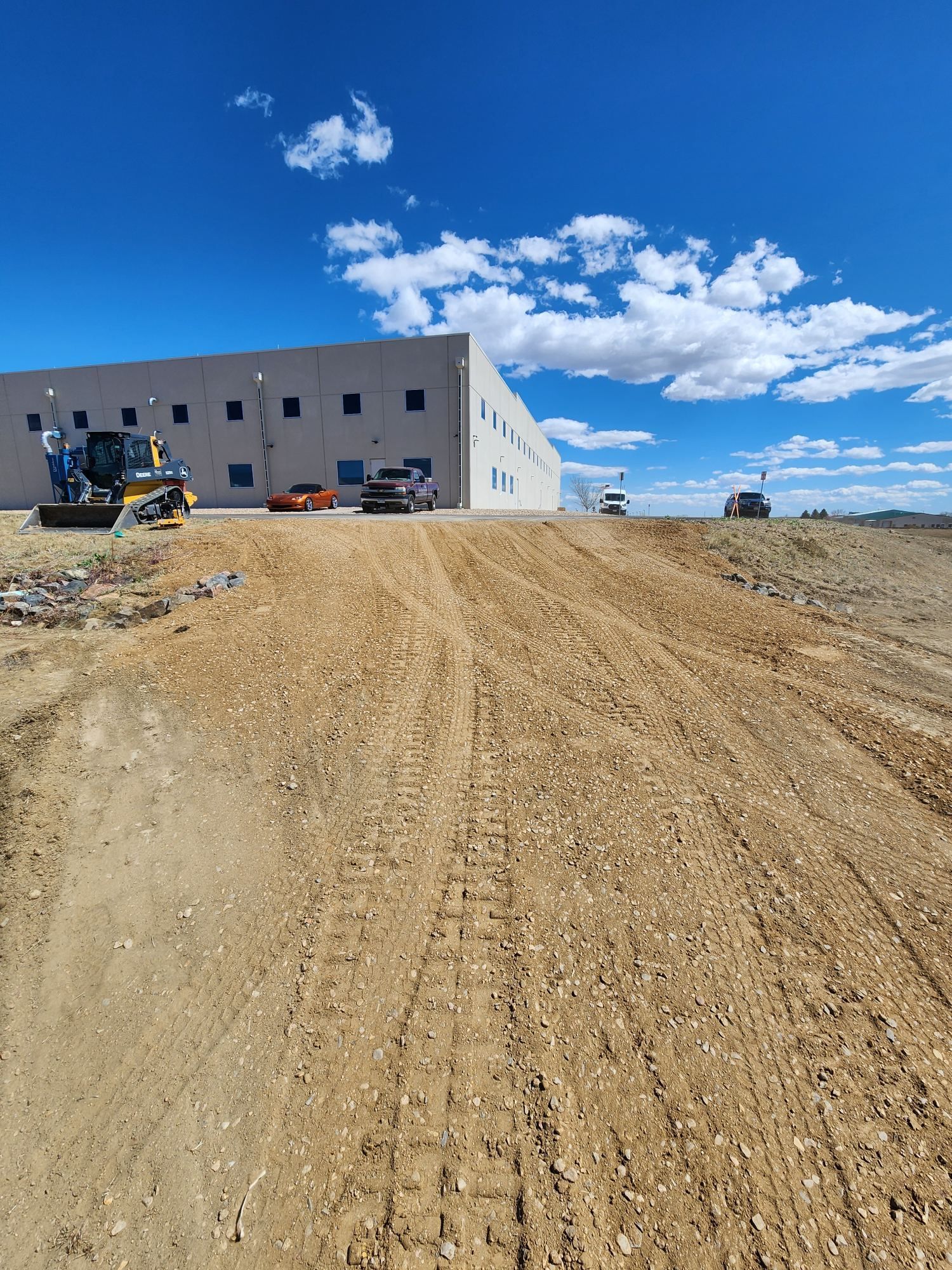 Construction site with a large building, dirt road, and excavator under a blue sky.