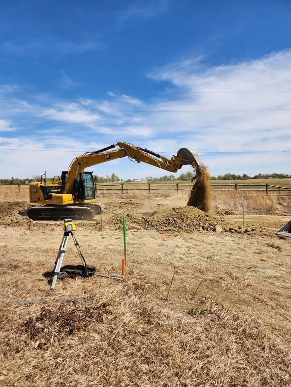 Yellow excavator on grass next to a black trailer, in front of a house.