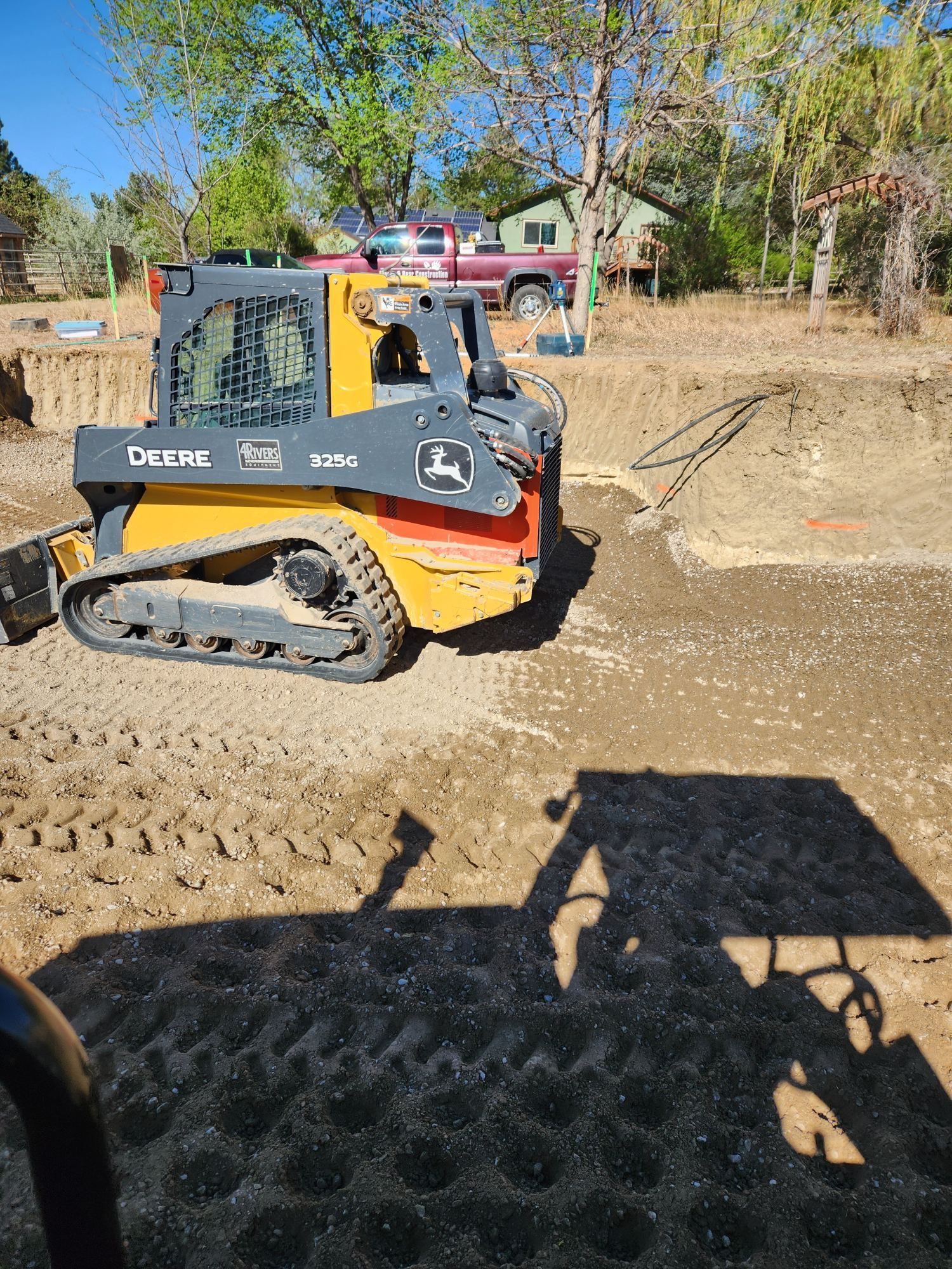 Compact track loader pushing dirt at a construction site, with trees and parked vehicles in the background.