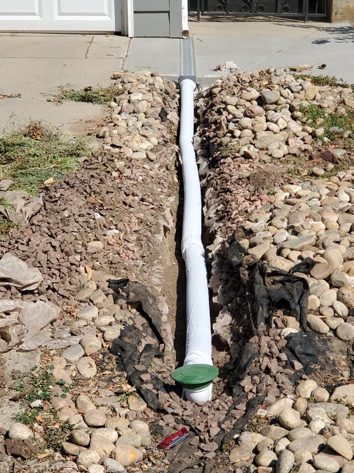 Trench with white drain pipe, green cap, and landscaping rocks near a garage door.