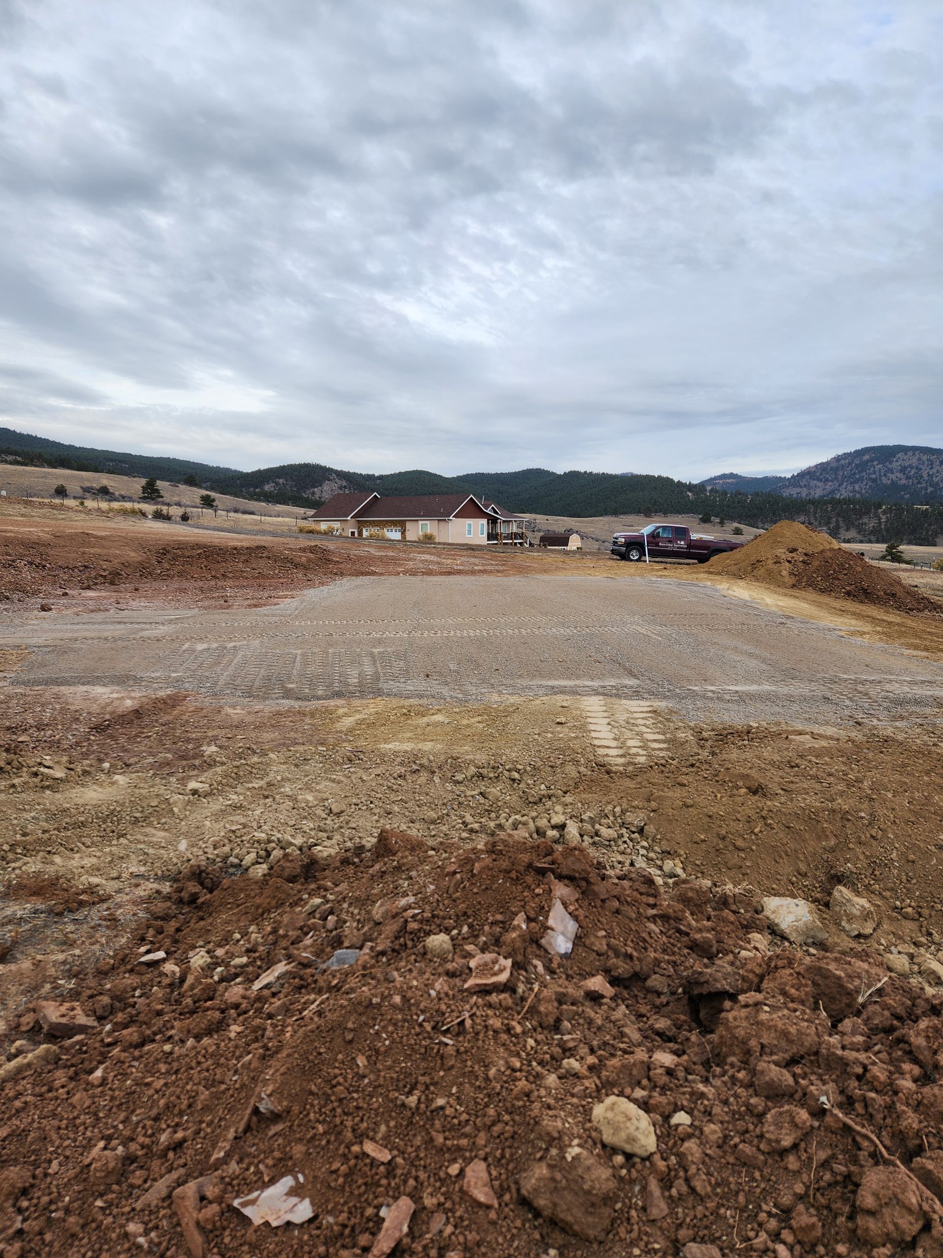 Gravel lot under cloudy sky, with muddy foreground and distant houses and hills.