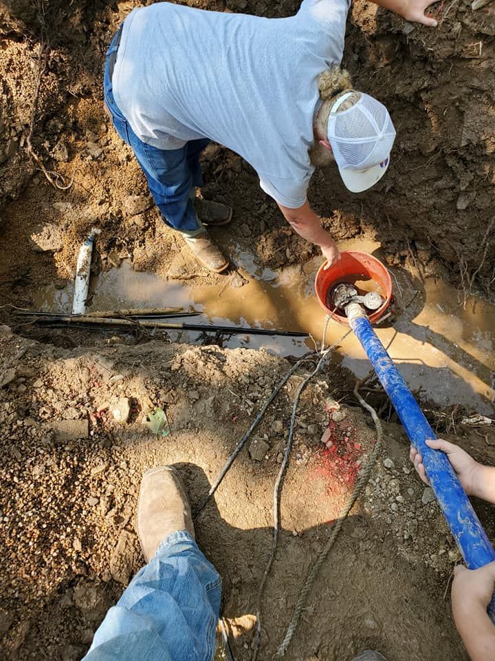 Two people repair a pipe in a muddy trench. One holds a bucket, while the other works on the pipe.