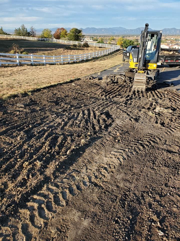 Mini excavator digging into dark soil next to a road, with a white fence, trees, and mountains in the background.