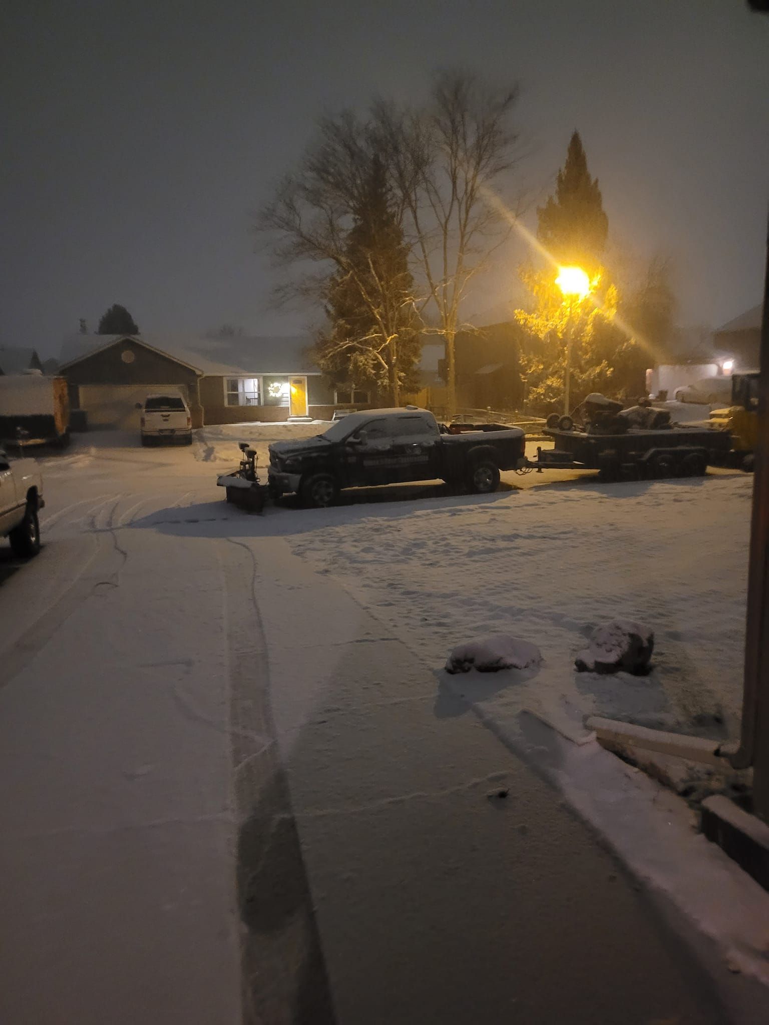 Snowy residential street at night, with a truck and trailer covered in snow near a brightly lit streetlight.