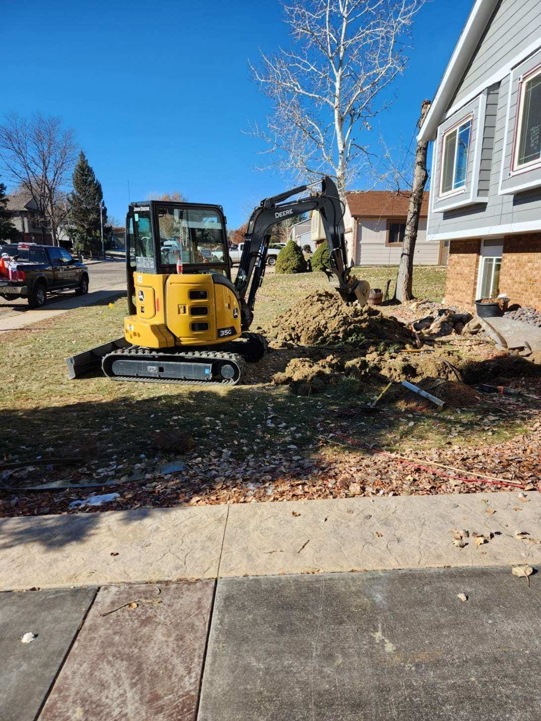 Yellow excavator digging in a residential yard next to a tree and house, under a clear blue sky.
