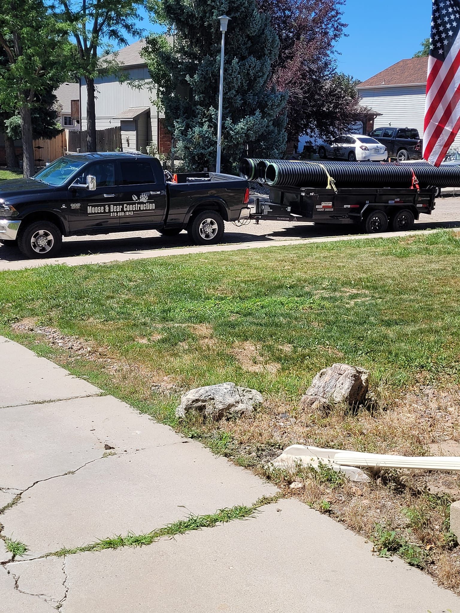 Black truck towing a trailer with dark-colored equipment on a sunny day near a residential area.
