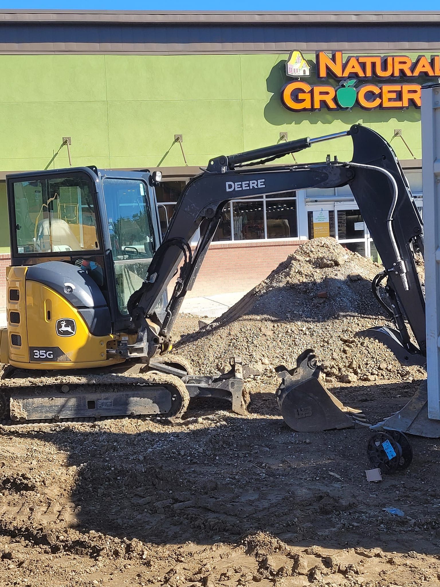 Yellow and black John Deere excavator in front of a Natural Grocers store, digging in gravel.