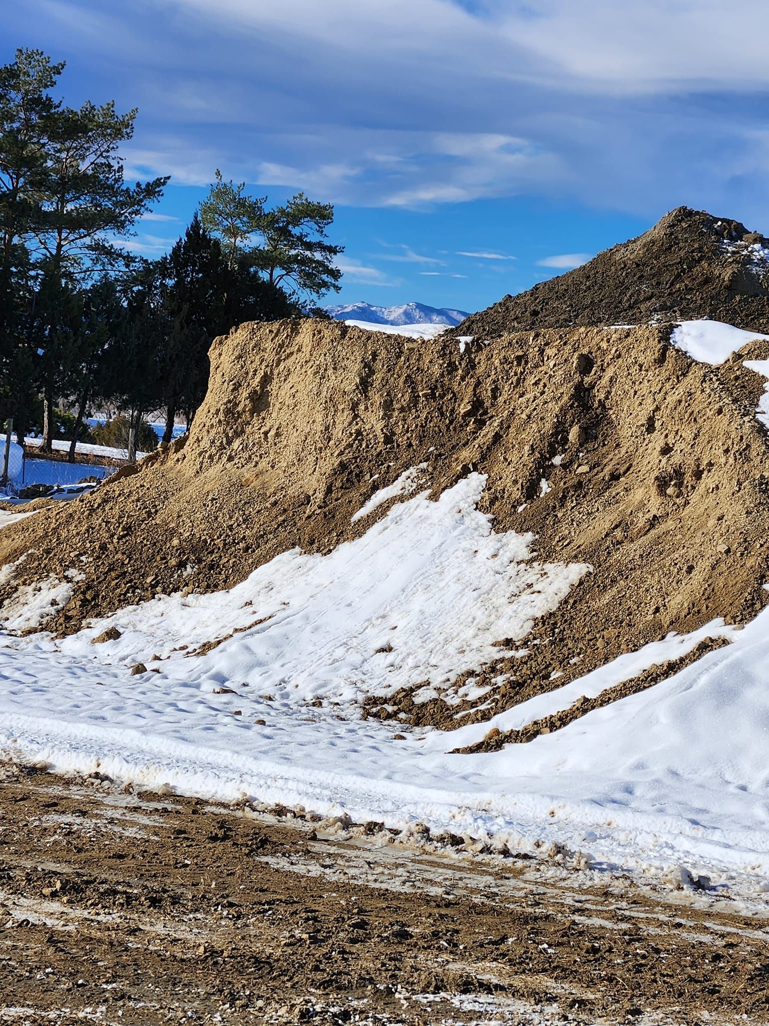 A pile of dirt and snow with trees in the background under a blue sky.