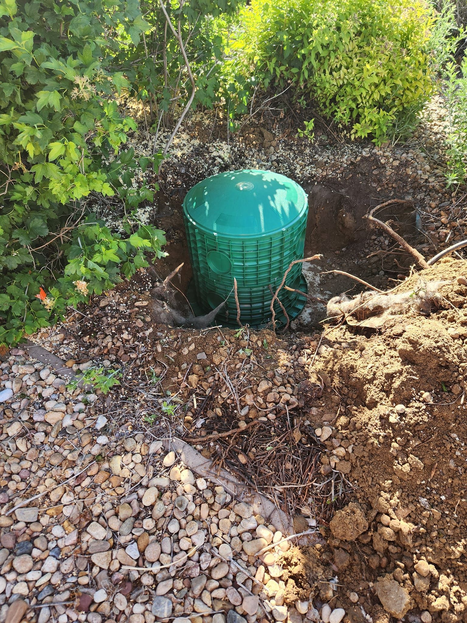 Green plastic cylinder partially buried in dirt, next to a rock bed and greenery.