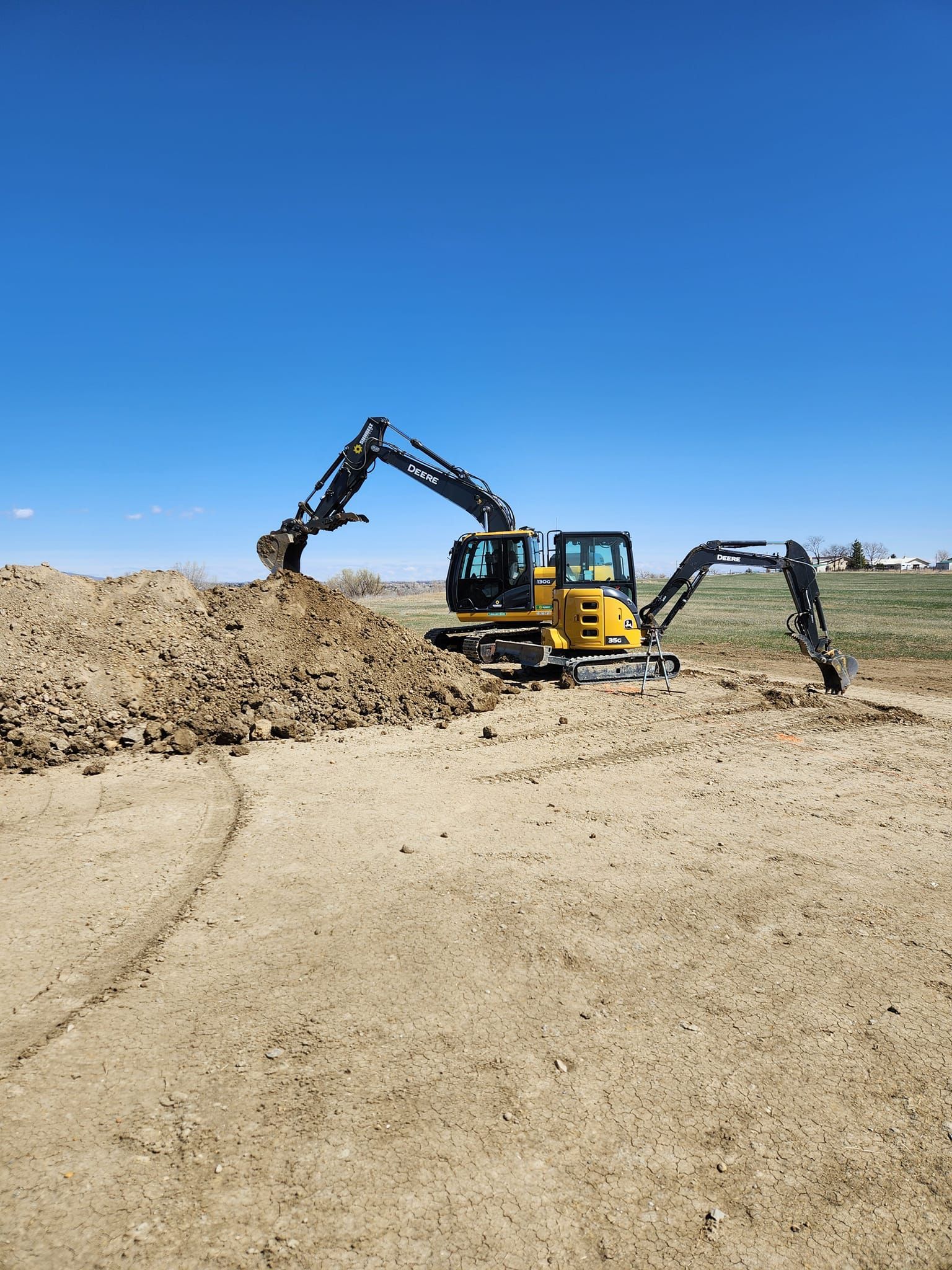 A yellow excavator with its arms extended on a dirt field under a blue sky, moving dirt.