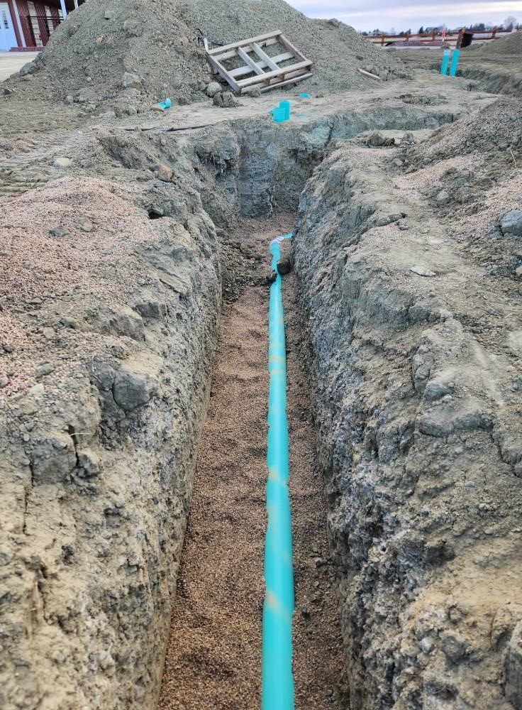 A trench with a turquoise pipe laid in gravel; dirt and a pile of soil in background.