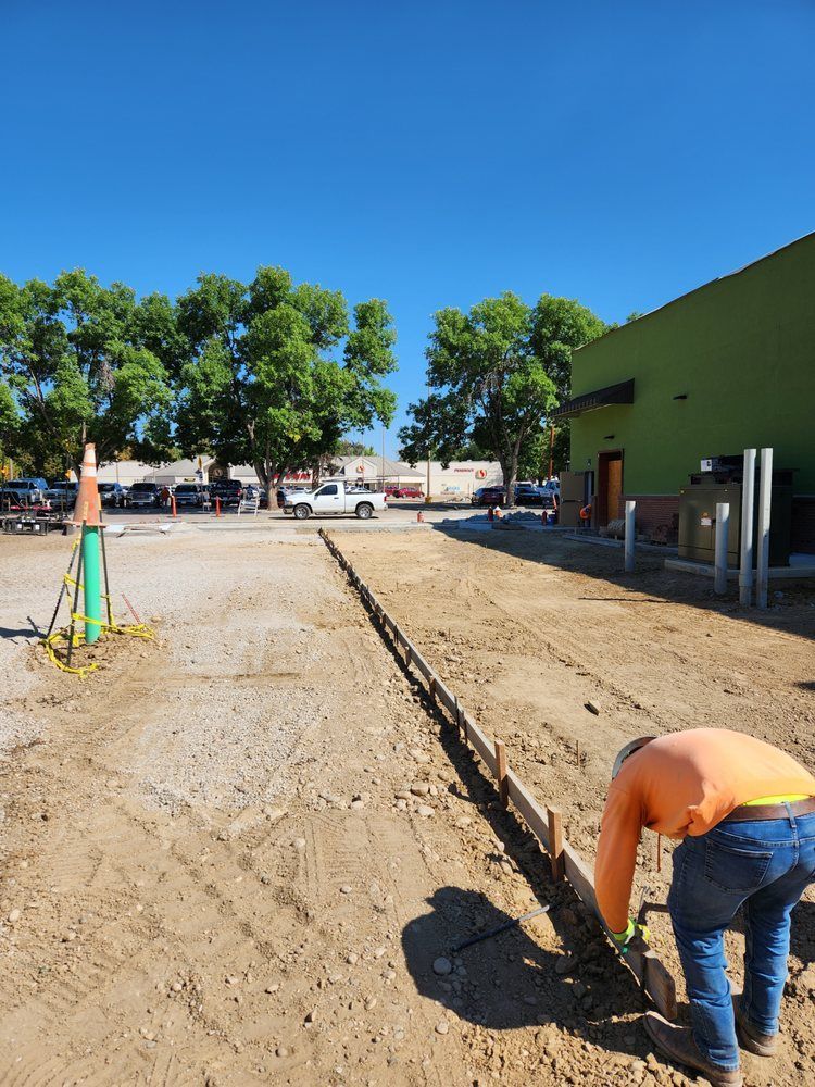 Construction worker working on a concrete form in a parking lot. Brown dirt, blue sky, green building, and trees.
