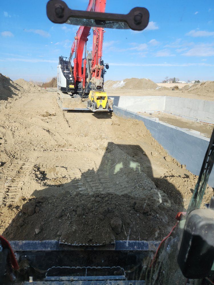 Construction site: Excavator working with a compactor on a dirt surface near a concrete wall, clear blue sky.