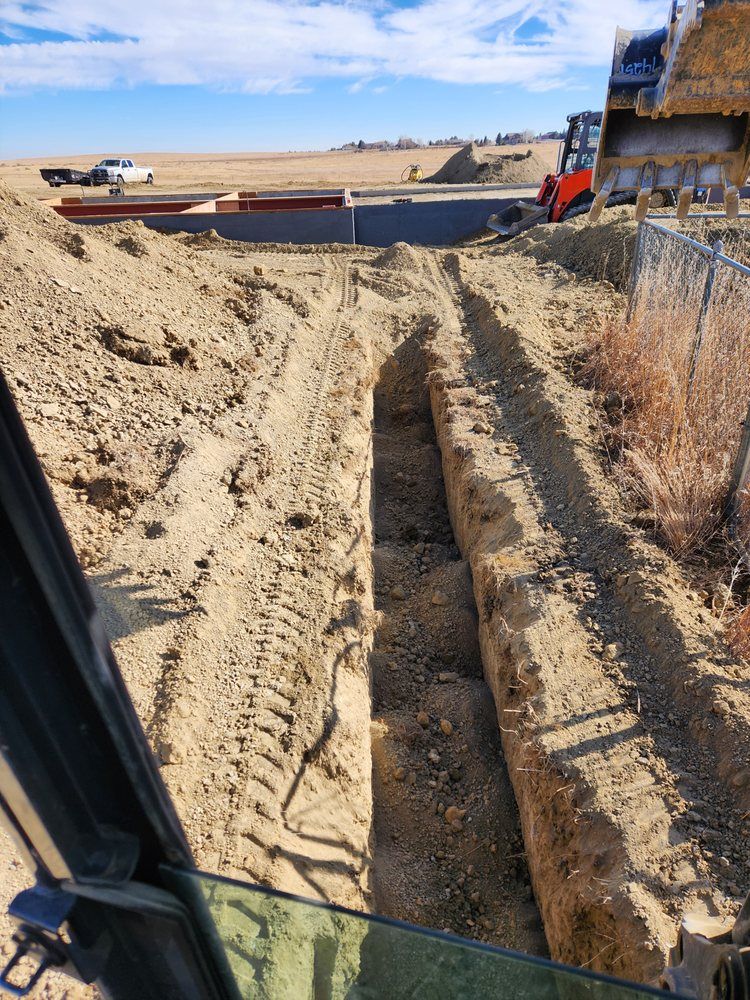 A construction site with a trench dug in the dirt, equipment in the background, clear sky.
