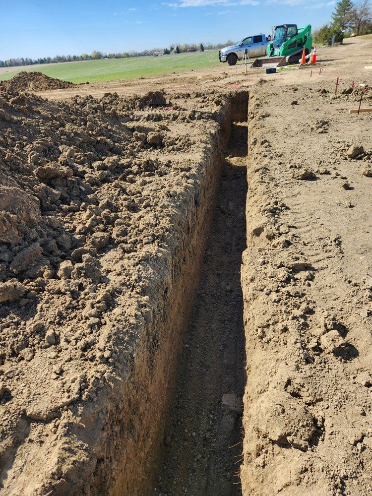Long narrow trench in dirt, with construction equipment in the background, likely for underground utilities.