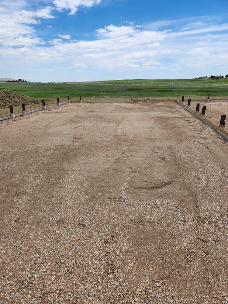 Gravel surface, possibly a shooting range, with wooden posts and green field in the background under a blue sky.