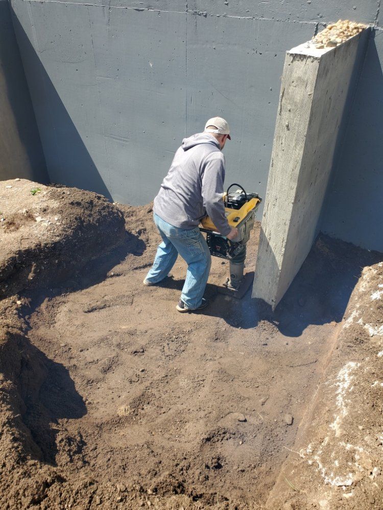 Person operating a plate compactor in an earthen trench next to a concrete structure.