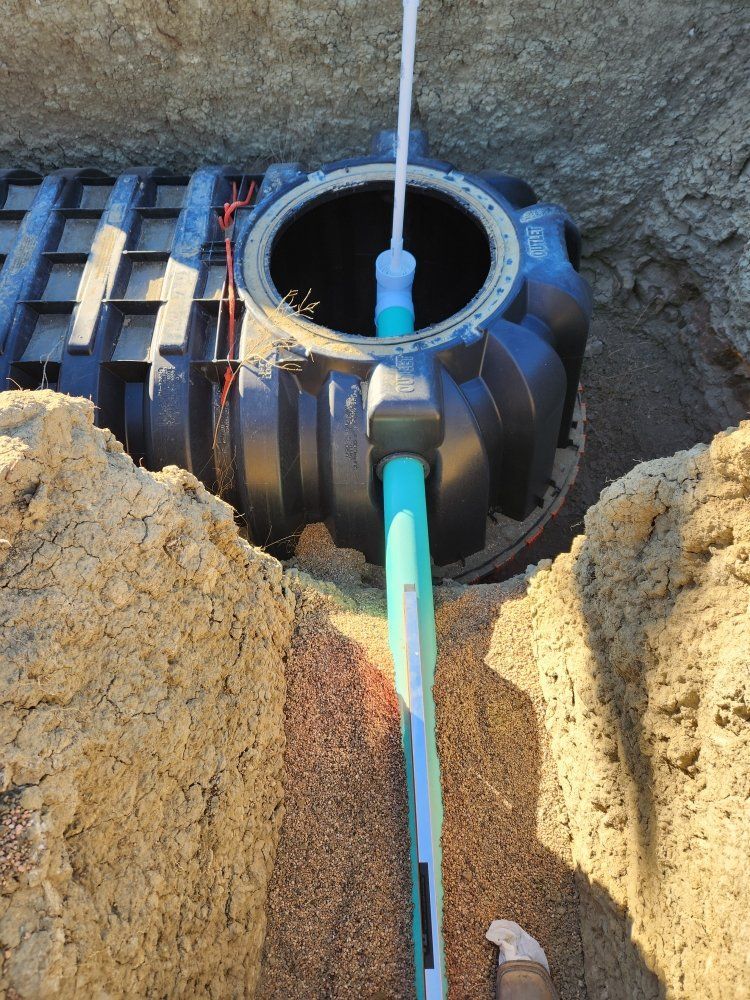 Black septic tank in a dirt trench, connected to a green pipe and topped with an open lid.