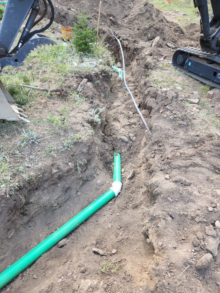 Trench dug in dirt with green pipes installed. Mini excavator and utility lines visible.