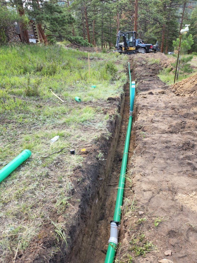 Trench with green pipes installed, construction site. Earthmoving in the background, surrounded by grass and trees.