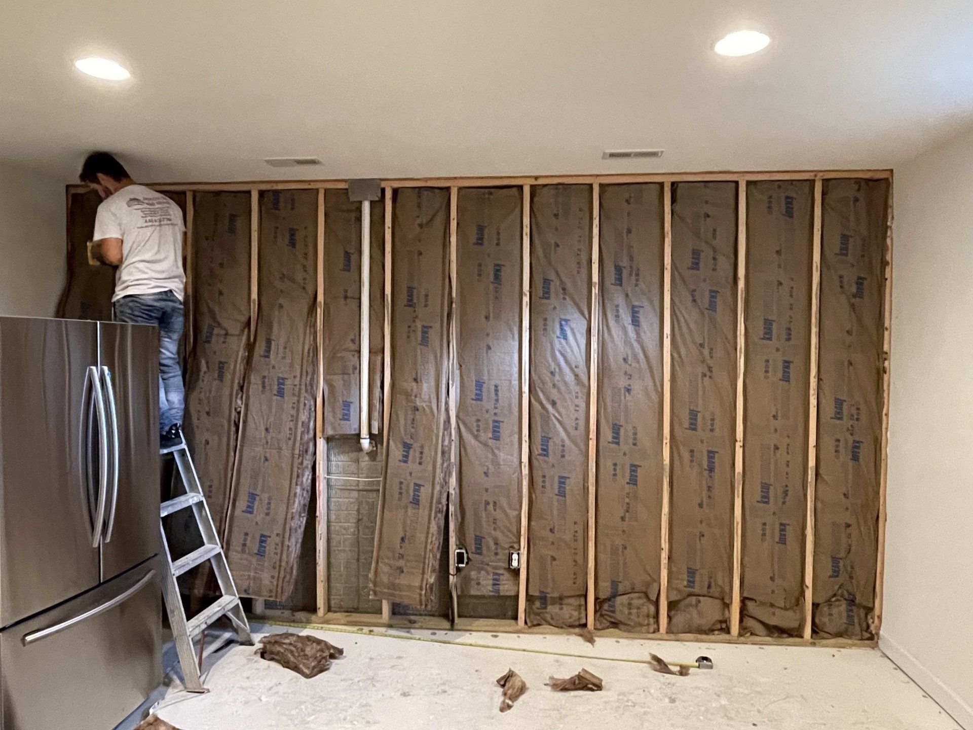 Person installing insulation between wall studs; a refrigerator is on the left.