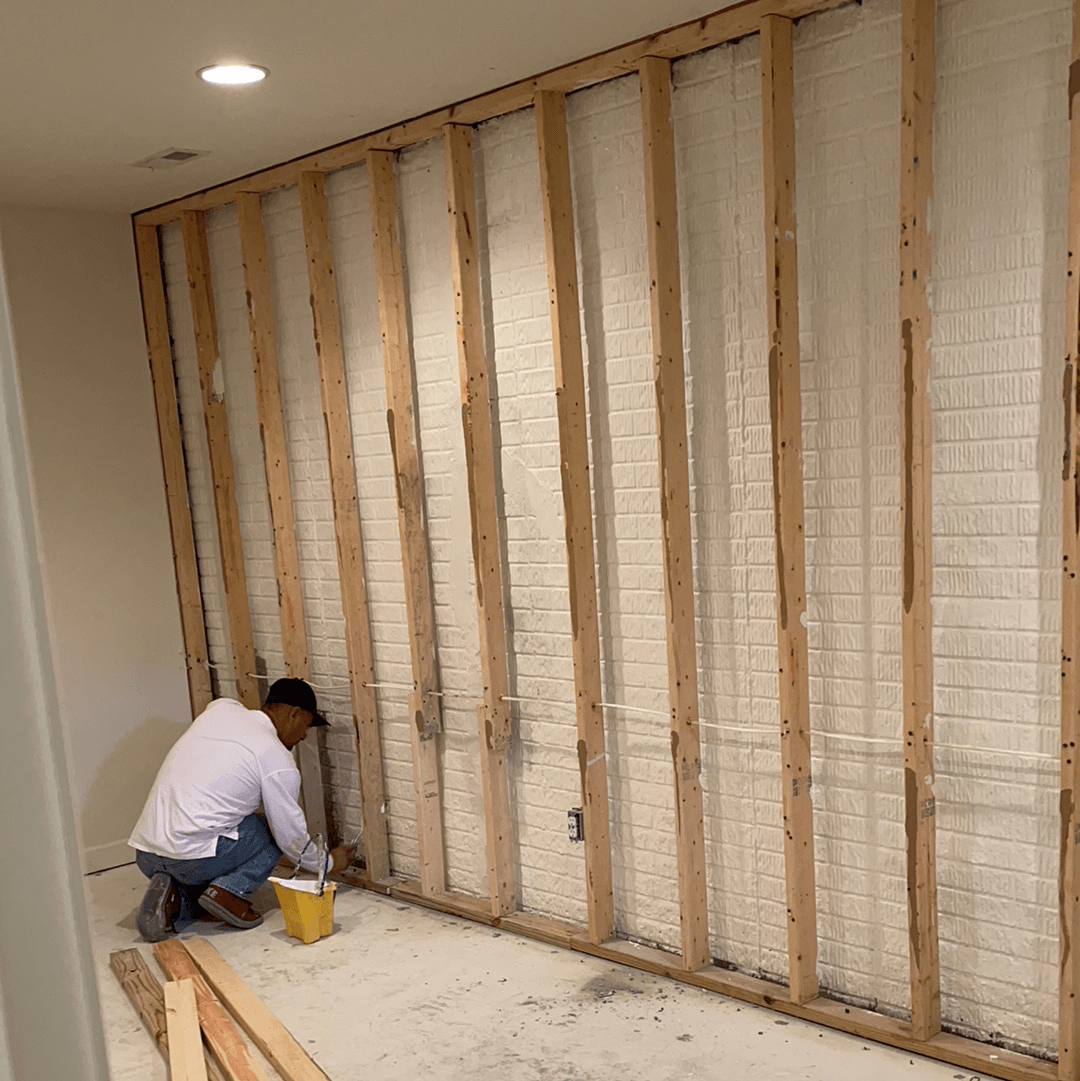 Man working on a framed wall, likely in a room under construction. Visible wooden studs and brick wall.