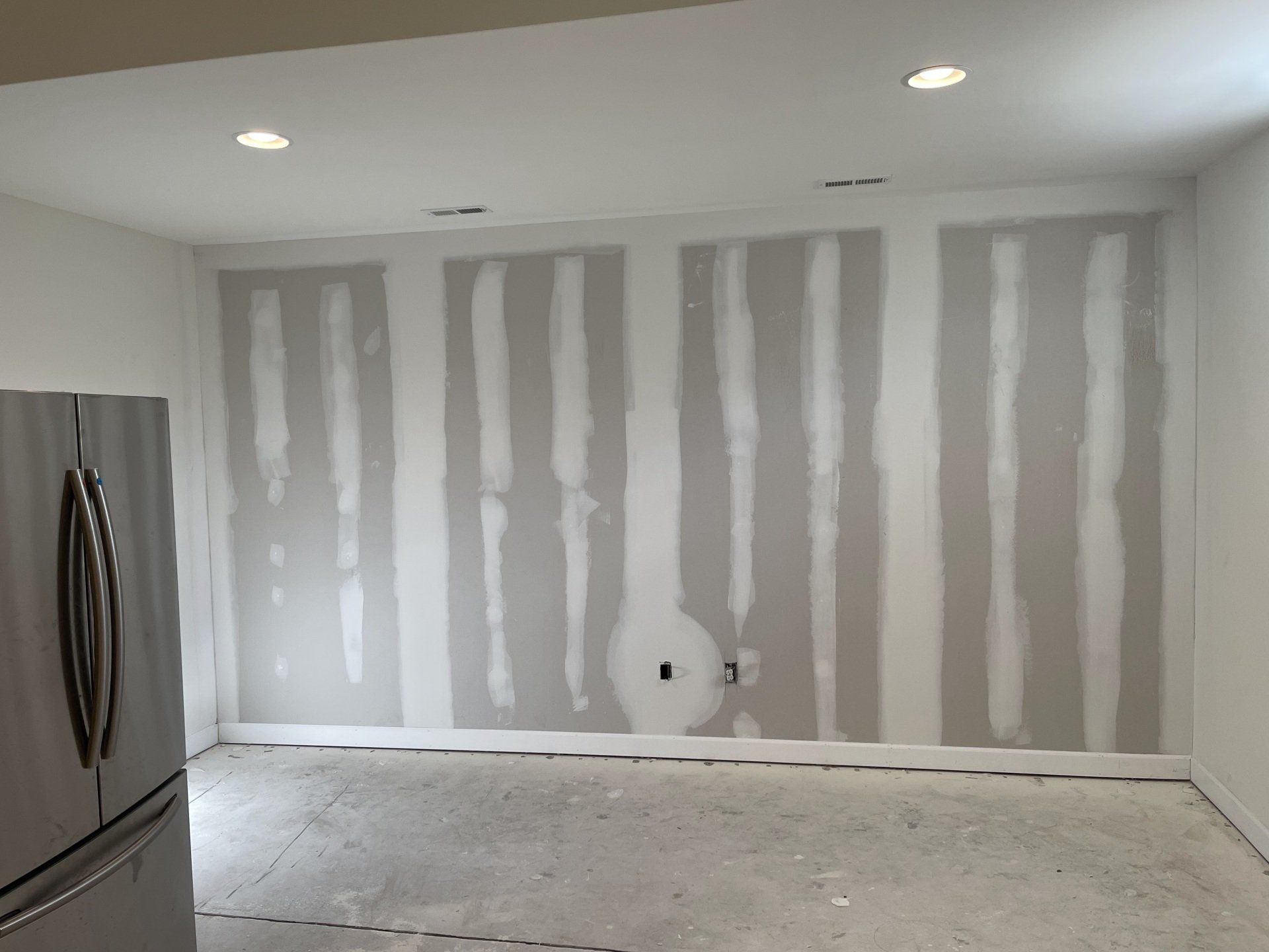 Room with unfinished drywall and partially finished seams; stainless steel refrigerator on left.