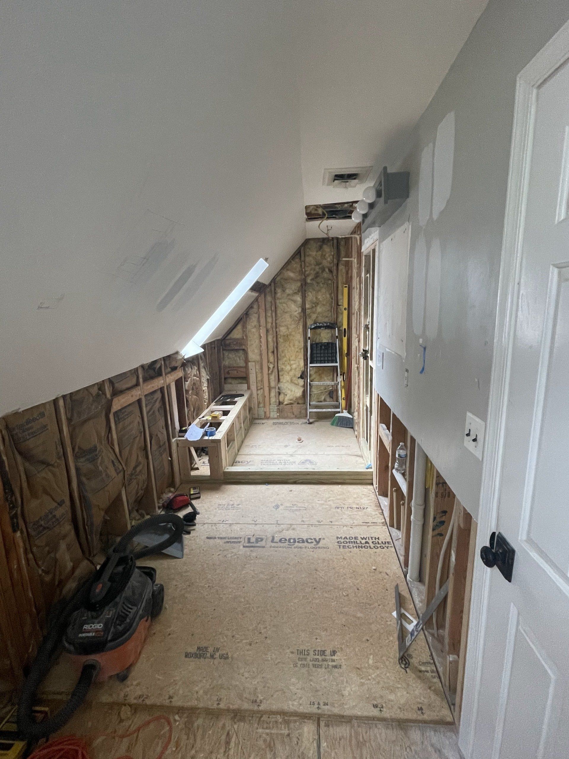 Attic renovation in progress; exposed studs, insulation, plywood flooring, and sloped ceiling.