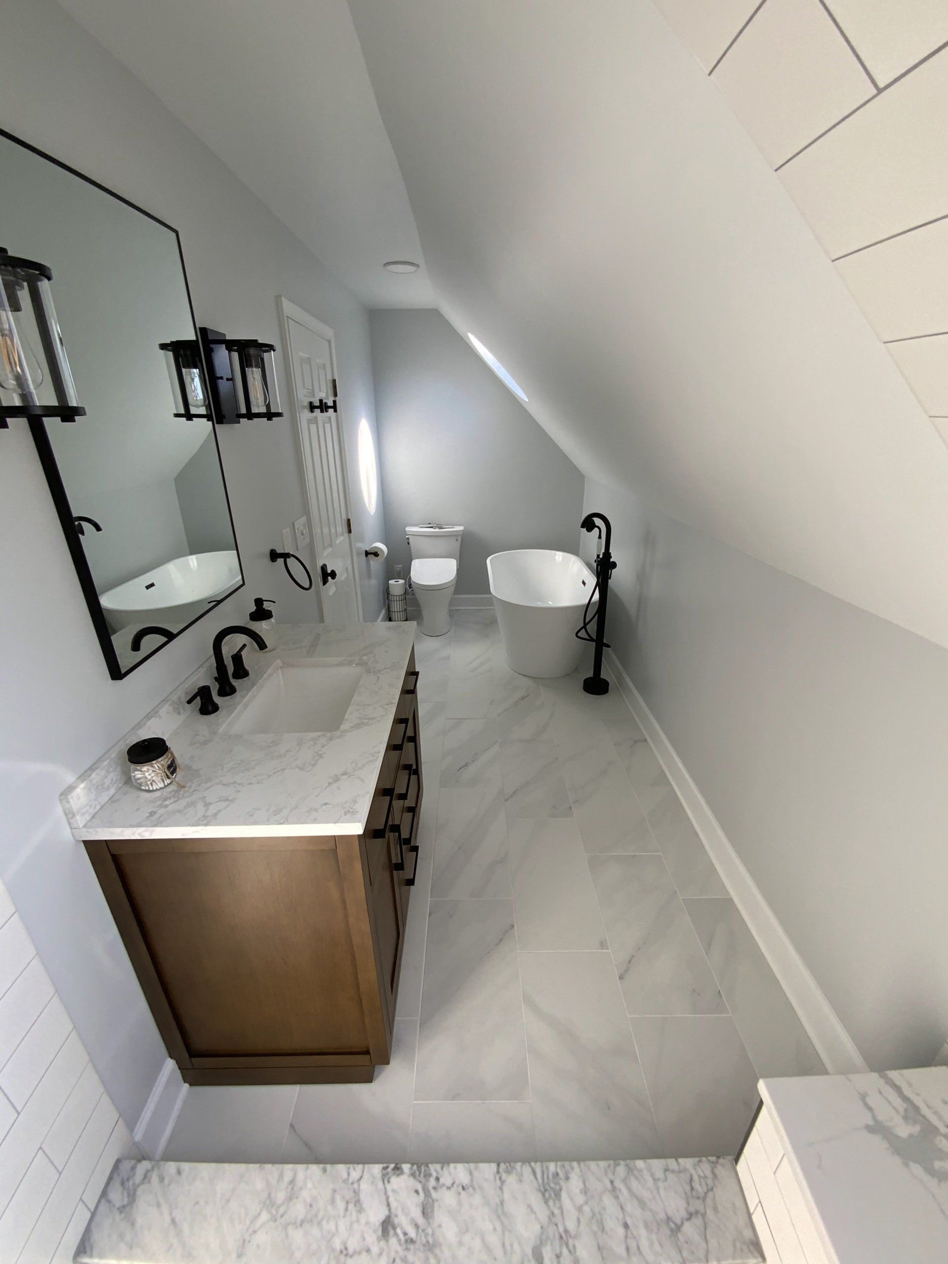 Bathroom with slanted ceiling, marble-look floor and countertop, white tub, brown vanity, and black fixtures.
