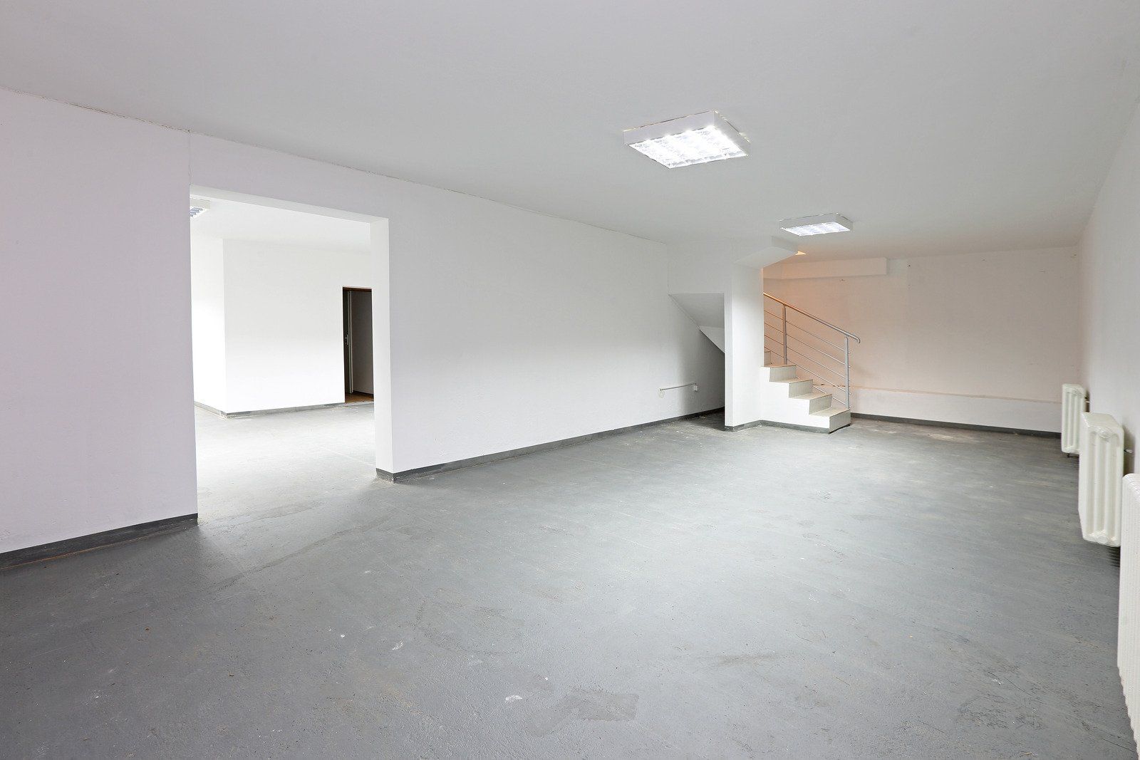 Empty white-walled room with gray floor; stairs to upper level, door to other room visible.