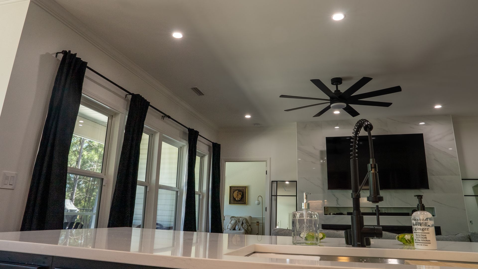 A modern kitchen with a white quartz island, black curtains, a large ceiling fan, and a flat-screen TV on a marble wall.